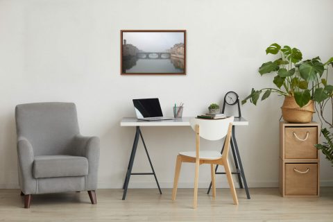A framed print of a foggy view of Ponte Santa Trinita in Florence, Italy, stands in a room with wooden decor. By Photographer Scott Allen Wilson.