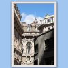 Wooden framed artistic print of a Genovese street scene, where grand adorned structures stand behind a curved, modern one and under a bright blue sky.