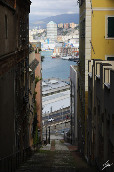 A narrow street in Genova, Italy, descends to the port of the city, while in the distance a beautiful cityscape opens up among the tall walls. Photo captured by Travel Photographer Scott Allen Wilson.
