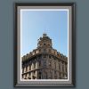 A black wooden framed piece of art of a gorgeous historical construction, filled with windows and decorative details, stands proud beneath a serene blue backdrop.