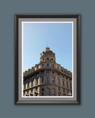 A black wooden framed piece of art of a gorgeous historical construction, filled with windows and decorative details, stands proud beneath a serene blue backdrop.