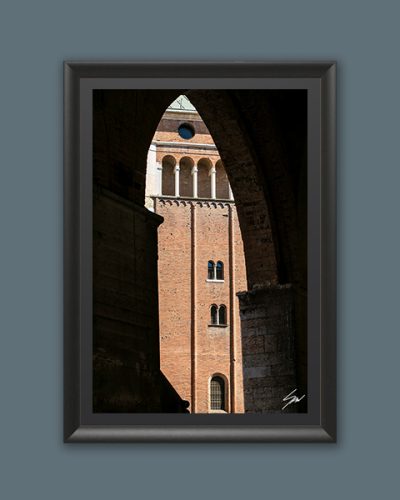 Beckoning by Scott Allen Wilson – framed fine art print of a sunlit medieval brick tower framed by a dark Gothic arch, Cremona, displayed in a black frame on a grey background