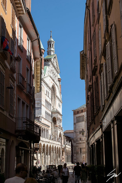 Ciao Cremona by Scott Allen Wilson – fine art photograph of a bustling narrow street in Cremona with tall residential buildings on either side leading to the ornate marble facade and green-domed spire of Cremona Cathedral against a vivid blue sky