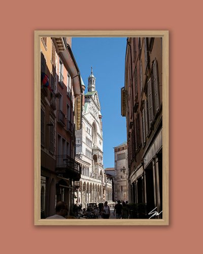 Ciao Cremona by Scott Allen Wilson – fine art print of a narrow Italian street leading to the ornate facade and spire of Cremona Cathedral under a clear blue sky, displayed in a natural wood frame on a terracotta background