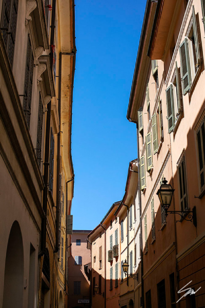 Scott Allen Wilson "Cremona Flow" fine art print showing a narrow Italian street canyon in Cremona, warm terracotta and ochre building facades rising on both sides toward a strip of vivid blue sky, green-shuttered windows and a wrought iron wall lantern visible on the right.