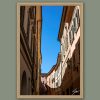 Scott Allen Wilson "Cremona Flow" fine art print in a natural wood frame against a sage green background, showing Cremona's historic street facades and blue sky.