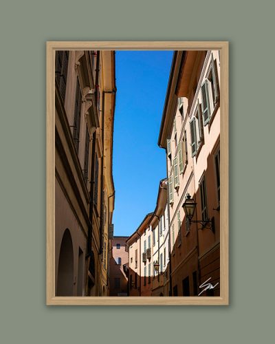 Scott Allen Wilson "Cremona Flow" fine art print in a natural wood frame against a sage green background, showing Cremona's historic street facades and blue sky.