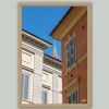Centered On You by Scott Allen Wilson – fine art print of two traditional Italian building facades framing a distant church spire under a vivid blue sky, Cremona, displayed in a natural wood frame on a grey background