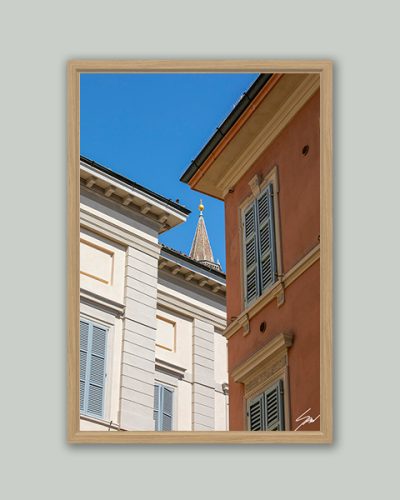 Centered On You by Scott Allen Wilson – fine art print of two traditional Italian building facades framing a distant church spire under a vivid blue sky, Cremona, displayed in a natural wood frame on a grey background