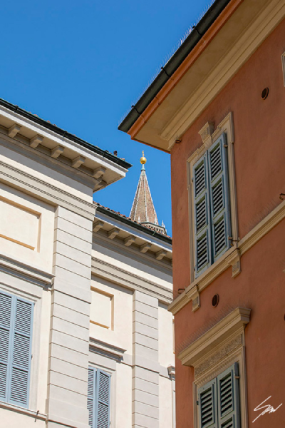 Centered On You by Scott Allen Wilson – fine art print of two traditional Italian building facades framing a distant church spire under a vivid blue sky, Cremona, displayed in a natural wood frame on a grey background