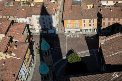 Fine art travel print of an aerial view of a historic Italian piazza with terracotta rooftops, colorful facades and a dramatic shadow sweeping across the square, The Source by Scott Allen Wilson, available in 8x12 and 16x24.