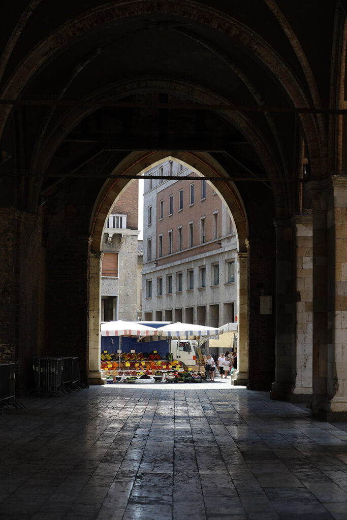 A market unfolds beyond the Gothic arches of Palazzo Gotico, blending everyday life with Piacenza's historic architecture.