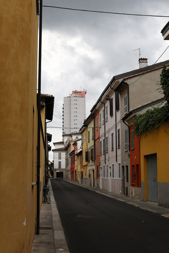 Narrow residential street in Piacenza lined with colorful historic buildings, with a modern tower rising in the distance beneath a cloudy sky.