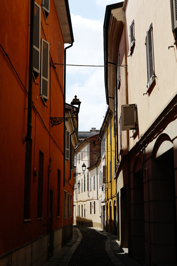 Narrow street in Piacenza lined with warm-toned facades, shuttered windows, and soft afternoon light filtering between historic buildings.