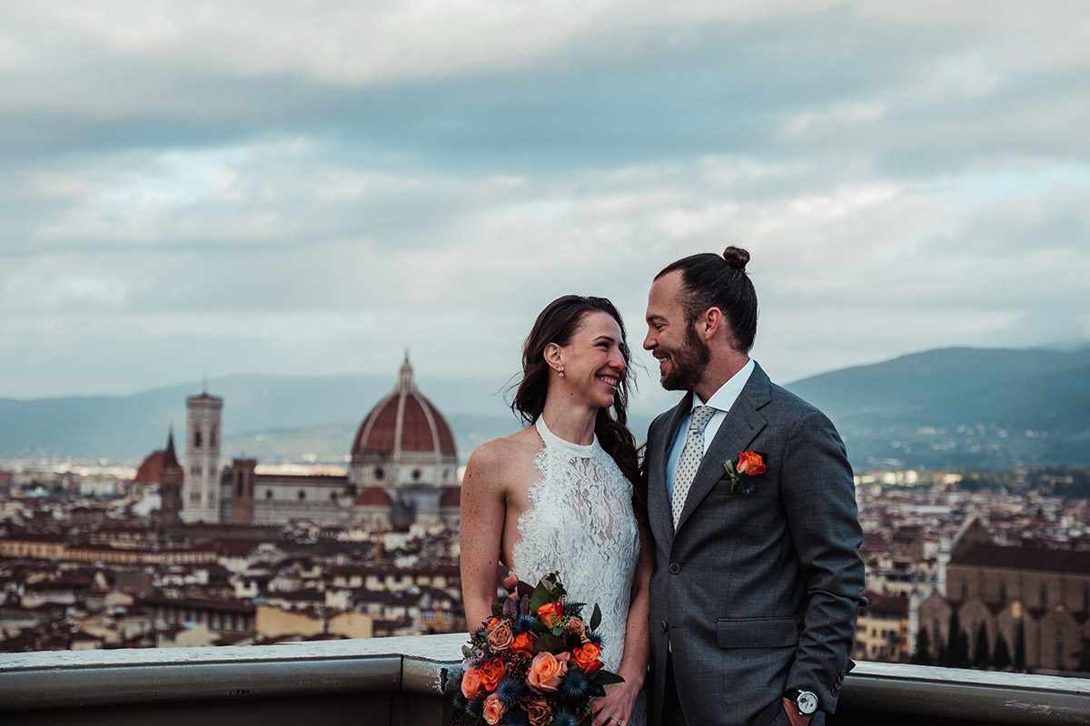 Gaby and Scott on a rooftop terrace with Florence Cathedral and the city skyline in the background, holding a bouquet of coral roses.