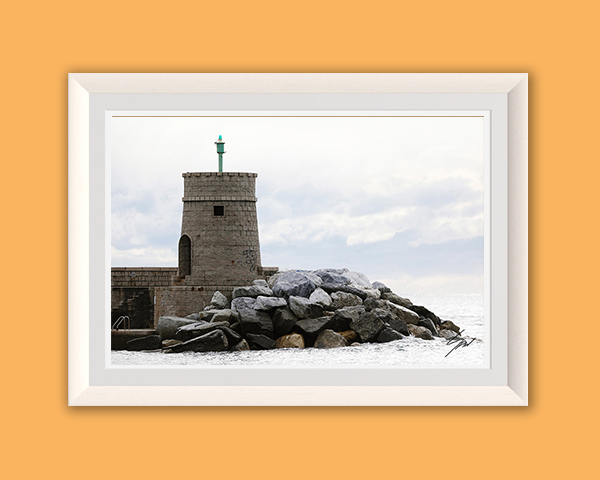 Fine art photograph of a historic stone breakwater tower standing against crashing waves and a cloudy Ligurian sky.