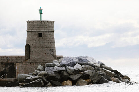 Fine art photograph of a historic stone breakwater tower standing against crashing waves and a cloudy Ligurian sky.