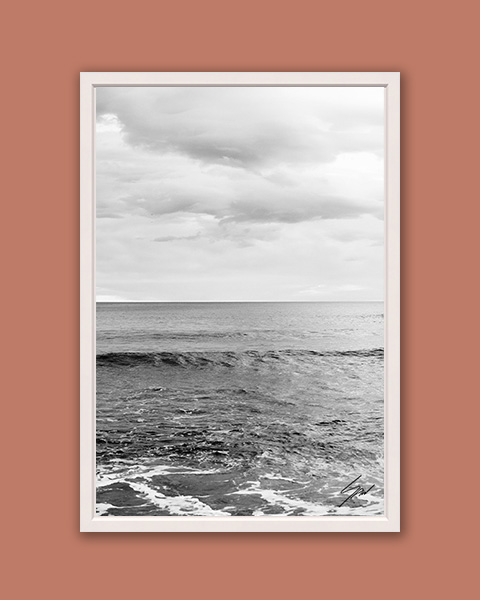 Black and white photograph of gentle waves meeting the horizon beneath a dramatic overcast sky.