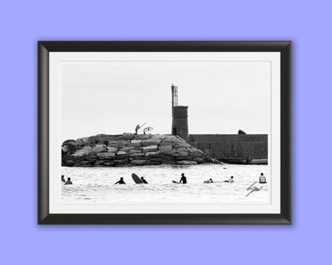 Coastal travel fine art print featuring surfers waiting beyond the break near a lighthouse in monochrome, creating elegant seaside wall art.