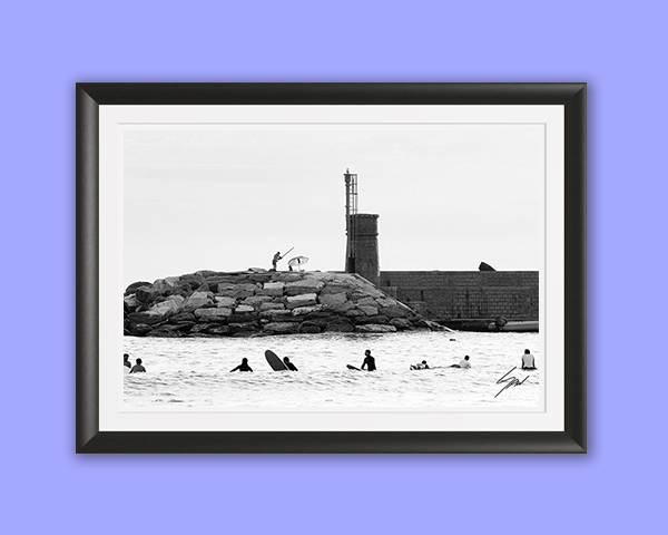 Coastal travel fine art print featuring surfers waiting beyond the break near a lighthouse in monochrome, creating elegant seaside wall art.