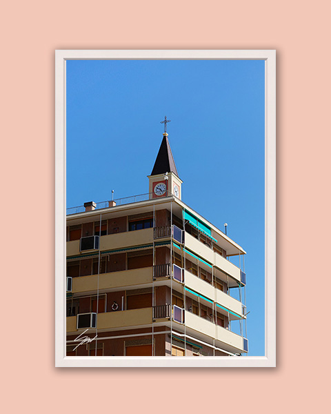 Fine art photograph capturing a church clock tower rising above a modern apartment building beneath a cloudless blue sky in Italy.