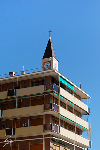 Fine art photograph capturing a church clock tower rising above a modern apartment building beneath a cloudless blue sky in Italy.