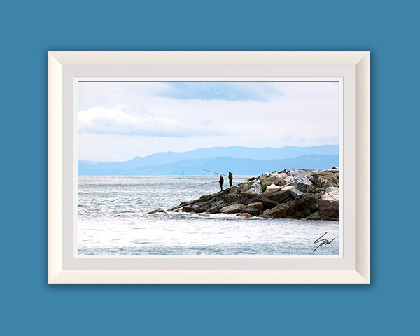Framed fine art print depicting a quiet coastal morning with fishermen on a stone jetty, calm sea, and layered mountains in the distance.