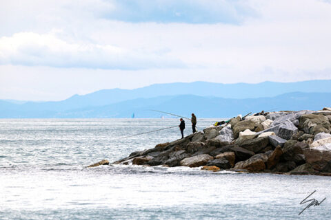 Framed fine art print depicting a quiet coastal morning with fishermen on a stone jetty, calm sea, and layered mountains in the distance.