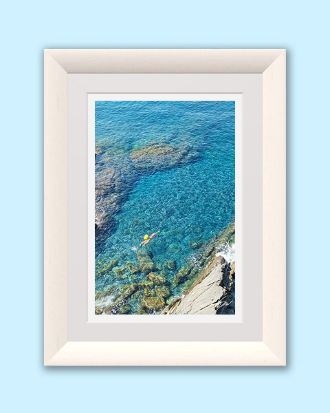 Overhead photograph of a lone swimmer wearing a yellow cap floating above crystal-clear turquoise water and rocky shoreline along the Ligurian coast.