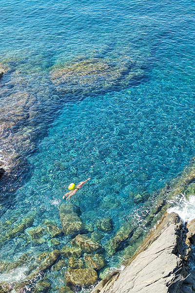 Overhead photograph of a lone swimmer wearing a yellow cap floating above crystal-clear turquoise water and rocky shoreline along the Ligurian coast.