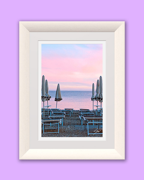 Rows of closed beach umbrellas facing a calm Mediterranean Sea beneath a soft pink and lavender sunset sky.