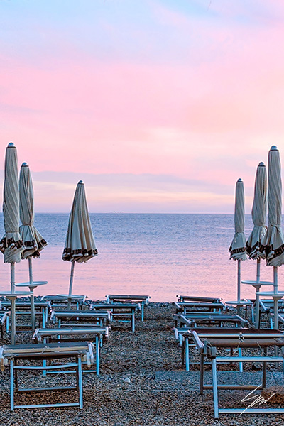 Rows of closed beach umbrellas facing a calm Mediterranean Sea beneath a soft pink and lavender sunset sky.