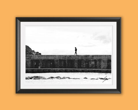Black and white coastal photography print capturing a lone walker crossing a historic breakwater on the Italian Riviera, perfect for contemporary wall decor.