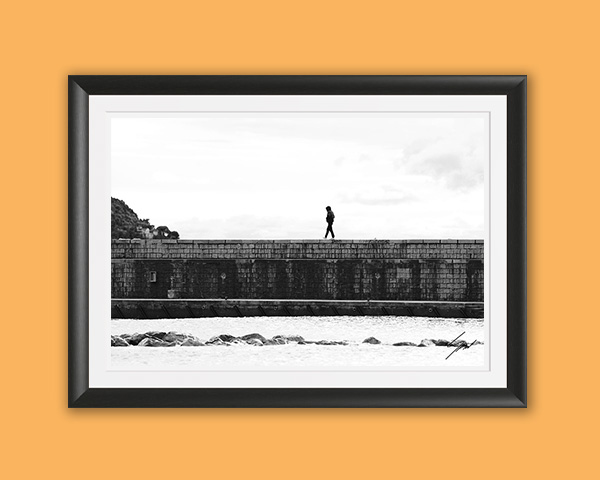 Black and white coastal photography print capturing a lone walker crossing a historic breakwater on the Italian Riviera, perfect for contemporary wall decor.