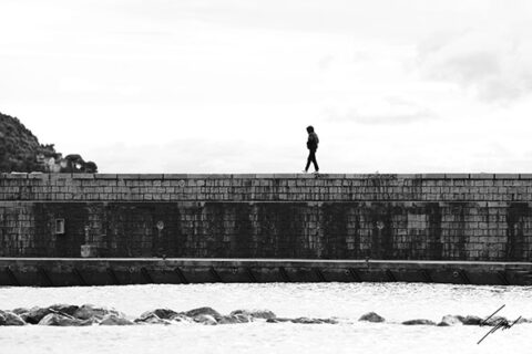 Black and white coastal photography print capturing a lone walker crossing a historic breakwater on the Italian Riviera, perfect for contemporary wall decor.