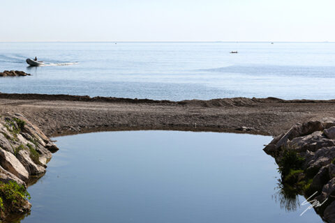 Peaceful coastal scene in Recco, Italy, where a still tidal pool mirrors the sky as a small boat glides across the Ligurian Sea.