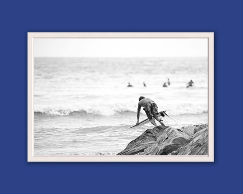 Black and white fine art photograph of a surfer climbing over seaside rocks before paddling into the waves on the Italian Riviera.