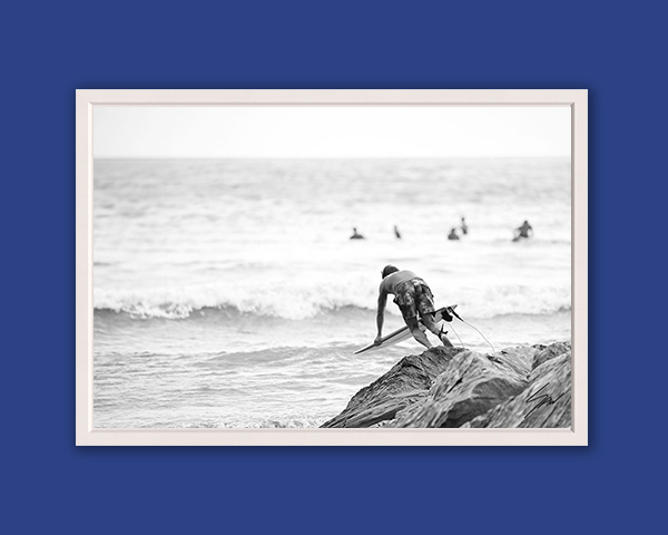 Black and white fine art photograph of a surfer climbing over seaside rocks before paddling into the waves on the Italian Riviera.