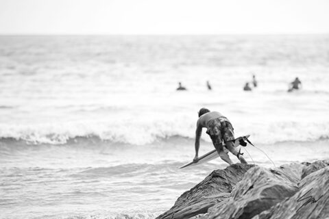 Black and white fine art photograph of a surfer climbing over seaside rocks before paddling into the waves on the Italian Riviera.