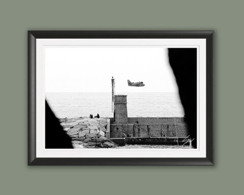Monochrome coastal photography print featuring a Coast Guard aircraft passing above a stone lighthouse, creating a striking blend of maritime history and aviation.