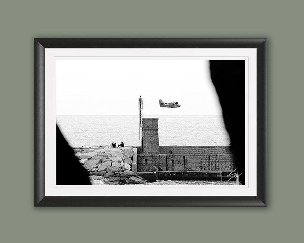 Monochrome coastal photography print featuring a Coast Guard aircraft passing above a stone lighthouse, creating a striking blend of maritime history and aviation.
