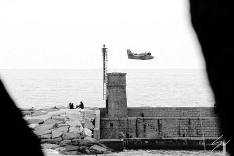 Monochrome coastal photography print featuring a Coast Guard aircraft passing above a stone lighthouse, creating a striking blend of maritime history and aviation.